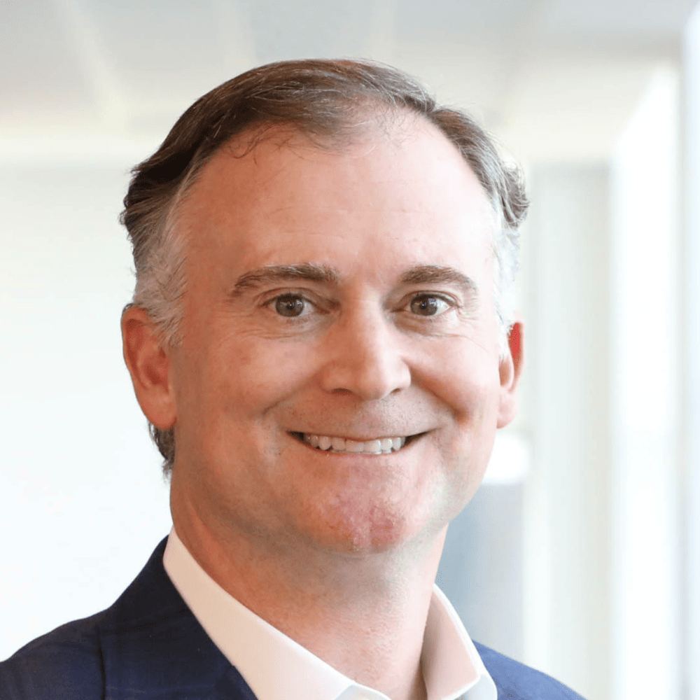 Headshot of Derek Van der Voort, from the shoulders up, smiling, in a charcoal blazer with short gray hair